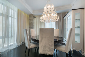 The interior of the living room in the apartment. An exclusive chandelier over a round marble table. White chairs with high backs.