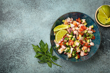 Fresh and vibrant peruvian seafood ceviche with shrimps, octopus, tomatoes, cucumber and parsley, served with lime in a bowl on a stone background from above, dish of Peru. Copy space