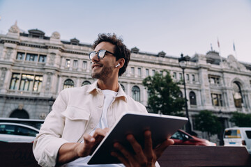 Smiling freelancer with smart glasses and tablet works outdoors, enjoying city views. Modern...