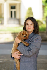 Beautiful woman in gray jacket and blue jeans holding cute maltipoo puppy in her hands. Dog and human relationship concept