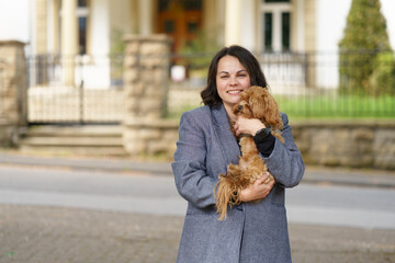 Beautiful woman in gray jacket and blue jeans holding cute maltipoo puppy in her hands. Dog and human relationship concept