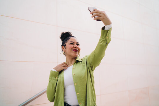 Young Hispanic woman confidently takes selfie, adjusting her necklace while smiling. Female appears to be capturing content for social media or a personal blog. Influencer and digital trends concept