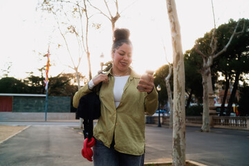 Smiling Hispanic woman walks outdoors, checking banking applications on smartphone. With fast 5G internet, female tech user effortlessly manages transactions and stays connected to digital services