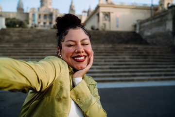 cheerful traveler smiles brightly while taking selfie, capturing joy against backdrop of historic architecture. Female relaxed pose and natural expression reflect a moment of happiness and adventure