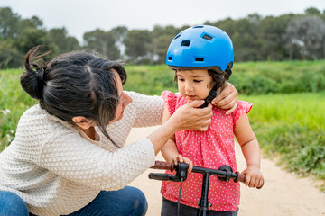 Young millennial mother helping toddler girl wearing a helmet learning to ride a bike outdoors. Concept of baby girl growing and developing, mother daughter bonding together outdoors parenting