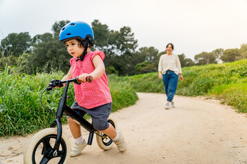 Fototapeta premium Toddler girl learning to ride a bike outdoors with her mother's help. Concept of baby girl growing and developing, mother daughter bonding together outdoors parenting