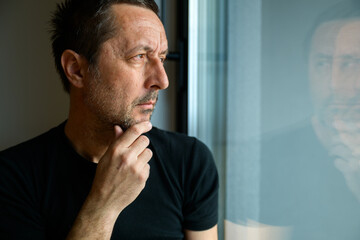 Fototapeta premium Serious caucasian mature adult male looking through window and thinking