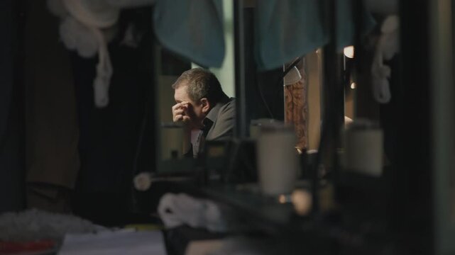 A man with blond  hair sits at table against mirror smearing paint over face. Man looks at reflection training facial expressions in dressing room.