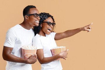 Happy black couple in 3D glasses eating popcorn, cheerful woman pointing at free space for advert...