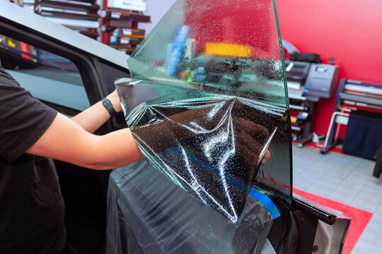 Worker applies tinted film to car window at workshop, showcasing meticulous detailing involved.