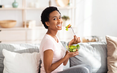 Healthy nutrition concept. Pretty black woman eating yummy vegetable salad on sofa at home,...