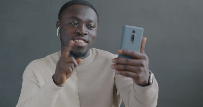 African American man making online video call with smartphone and earphones talking gesturing on gray color background. People and communication concept.