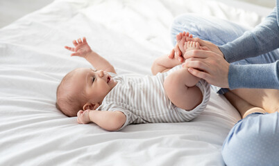 Caring Mother Making Gymnastics To Her Newborn Baby On Bed At Home, Unrecognizable Woman Helping...