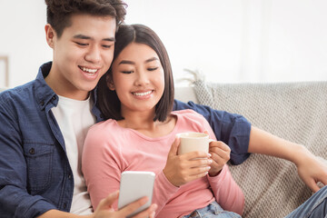 Obraz premium Korean Couple Using Smartphone Relaxing Sitting On Sofa At Home On Weekend. Cellphone Users Browsing Internet, Gadget Lifestyle, Mobile Application Concept. Empty Space