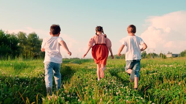 Children team runs through green meadow, dreaming of flight. Children boy girl, friends happily run through grassy meadow with flowers. Happy team of children play outdoors on green field against sky
