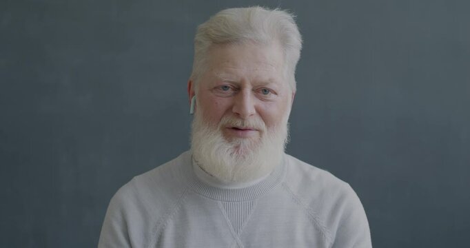 Portrait of senior man making online video call talking and gesturing looking at camera using wireless earphones on black color background.