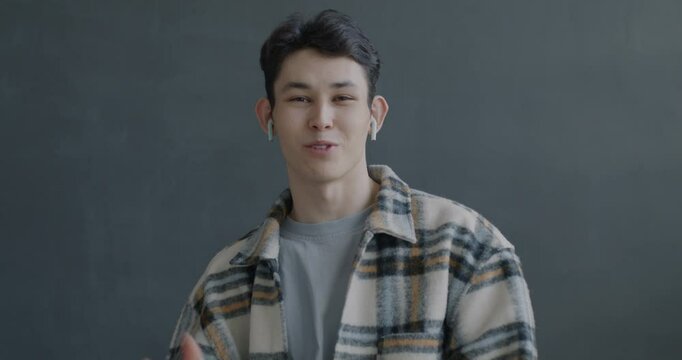 Portrait of joyful Asian man making online video call using earphones talking looking at camera on gray color background. People and communication concept.