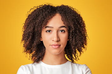 Human emotions and stern look of person. Serious, calm millennial african american lady with curly hair in white t-shirt looking at camera, isolated on yellow background, close up, free space