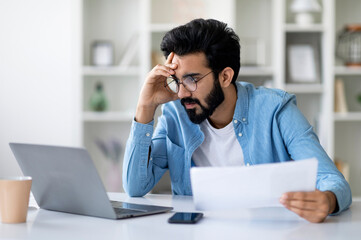 Stressed Young Indian Man Looking At Laptop Screen And Holding Documents, Focused Millennial...