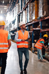 Warehouse manager walking and using tablet with workers checking inventory in background