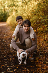 Fototapeta premium Front view of father and young son in warm clothes on autumn forest path walking with dog, smiling and looking at camera. Concept of family, bonding, fatherhood, parenting, leisure.