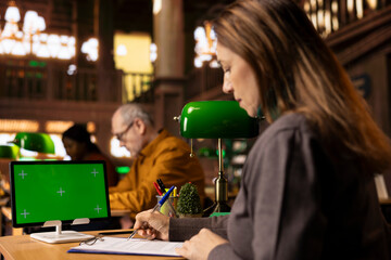 Wise adult learner focused taking notes for research next to chroma key, gathering information and sources for citations. Mature woman working on her diploma paperwork in a public library.