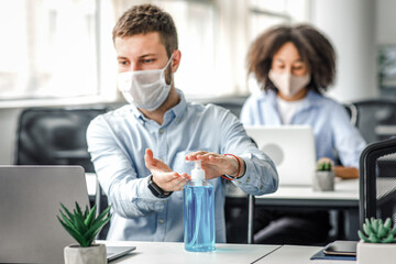 Obraz premium Protection at coronavirus pandemic in office. Young man in protective mask works at laptop and disinfects his hands with antiseptic
