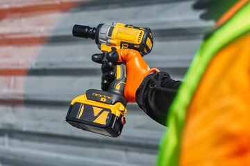Close-up construction worker wearing high visibility safety vest and protective gloves, holding a cordless power drill. Industrial work and safety gear with construction tools.
