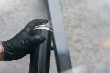 Close-up of a hand wearing a black glove spraying paint from a black aerosol can onto a metal surface.