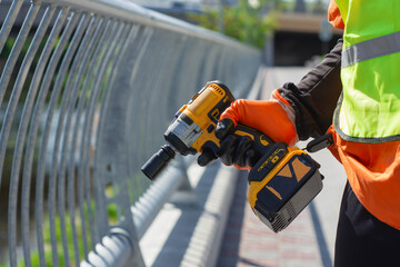 A worker in a safety vest and orange gloves holds a power drill near a metal railing on a...