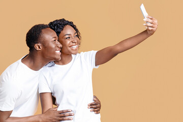 Cheerful african american couple taking selfie on smartphone over yellow studio background, copy space. Happy young black man and woman lovers taking photos while bonding, using mobile phone