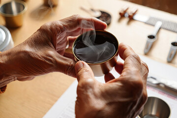 Close-up of elderly person's hands with dark skin holding a small metallic cup of steaming hot black coffee during a cupping or tasting session with blurred coffee tools on a wooden table under natura
