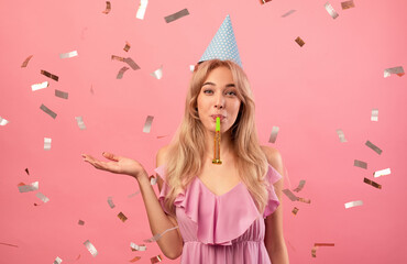Gorgeous young woman in birthday cap blowing party horn on pink studio background with falling...