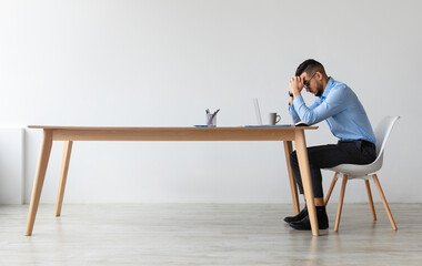 Profile side view of young Middle Eastern man suffering from headache at workplace, having problems thinking about troubles. Stressed guy in glasses sitting at desk using computer, free copy space