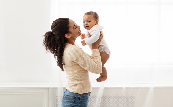 Mom's Love. Young Black Mother Holding Baby Toddler Playing And Cuddling With Little Infant Son Standing Near Window Indoor. Motherhood Happiness And Child Care, Maternity Leave Lifestyle. Side View