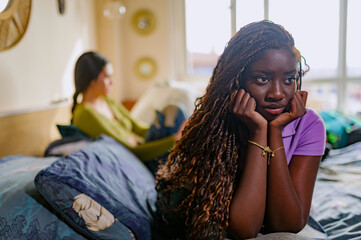 Sad woman with long braided hair resting head in hands while her partner sits on bed in background,...
