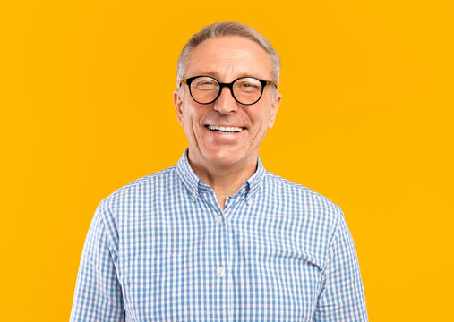 Portrait of senior male smiling wearing glasses and casual checkered shirt posing standing isolated over yellow orange studio background. Happy excited adult person looking at camera, free copy space