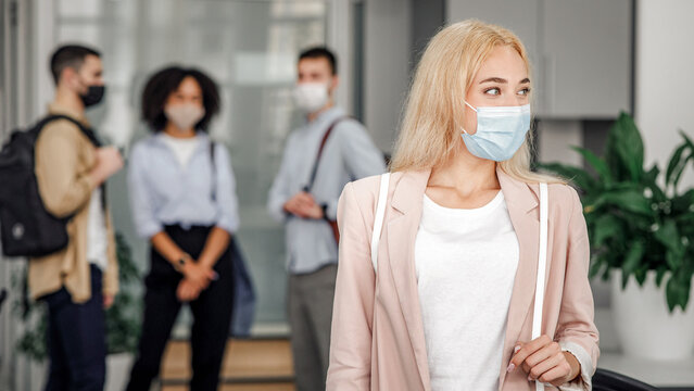 Workers in modern office hallway return to work after quarantine. Focus on beautiful blonde woman in business suit and protective mask looks to the side and blurred background, empty space