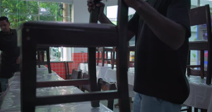 Restaurant workers stacking chairs on tables at end of day, preparing dining room for cleaning in casual indoor setting with natural window light