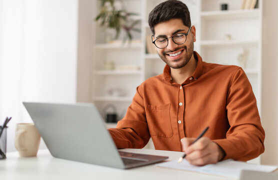 Portrait of young smiling Arabic man in glasses using laptop sitting at desk, writing in notebook. Happy guy working remotely, watching webinar training studying online, taking notes signing documents