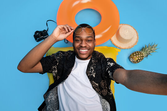 Top view of smiling black guy in summertime outfit lying on lilo, taking selfie on beach vacation, blue studio background. Positive young man making photo of himself, enjoying summer holidays