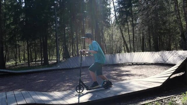 A 12 year old boy in a cap and shorts rides a scooter on a pump track in Borovsky Park in Moscow in the spring.	