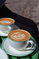 Two white elegant cups of lattes with latte art on a green table. Morning coffee break on cafe terrace. Decorated cappuccino with artful foam in a mug