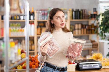 Young woman buyer chooses raw sausages and pork meat in grocery store