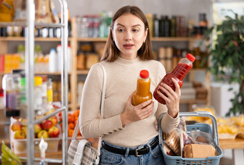While shopping in grocery store, girl buyer choosing juice. Client look package of fruit drink,...