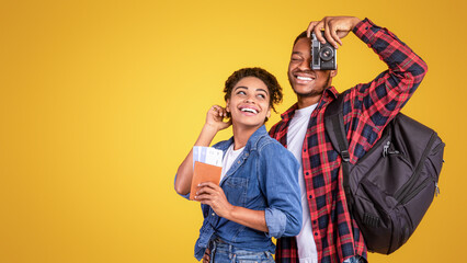 Obraz premium Travel. Cheerful African Tourists Couple With Photo Camera And Luggage Standing Ready For Vacation Posing On Yellow Studio Background