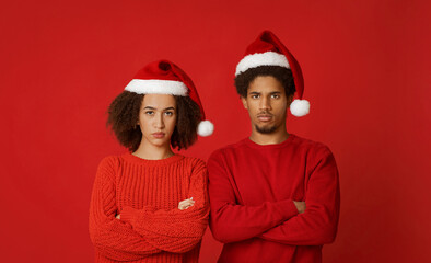 Fototapeta premium Unlucky celebration of christmas or new year. Sad and upset african american young guy and woman in santa claus hats with arms crossed on their chest, looks at camera, isolated on red background