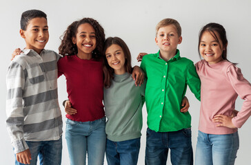 Cheerful Multicultural Preteen Kids Standing Hugging And Smiling To Camera Posing Together Over Gray Studio Background. Shot Of Mixed School Boys And Girls Group. Friendship Concept