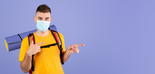 Traveling during coronavirus epidemic concept. Young man with camping equipment wearing medical mask, pointing aside over blue studio background. Millennial tourist staying safe during viral pandemic
