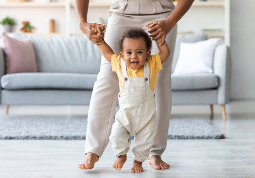 Adorable black infant boy making first steps at home with mother's help. Happy smiling cute african american toddler baby holding mom's hands while walking in living room, closeup with copy space